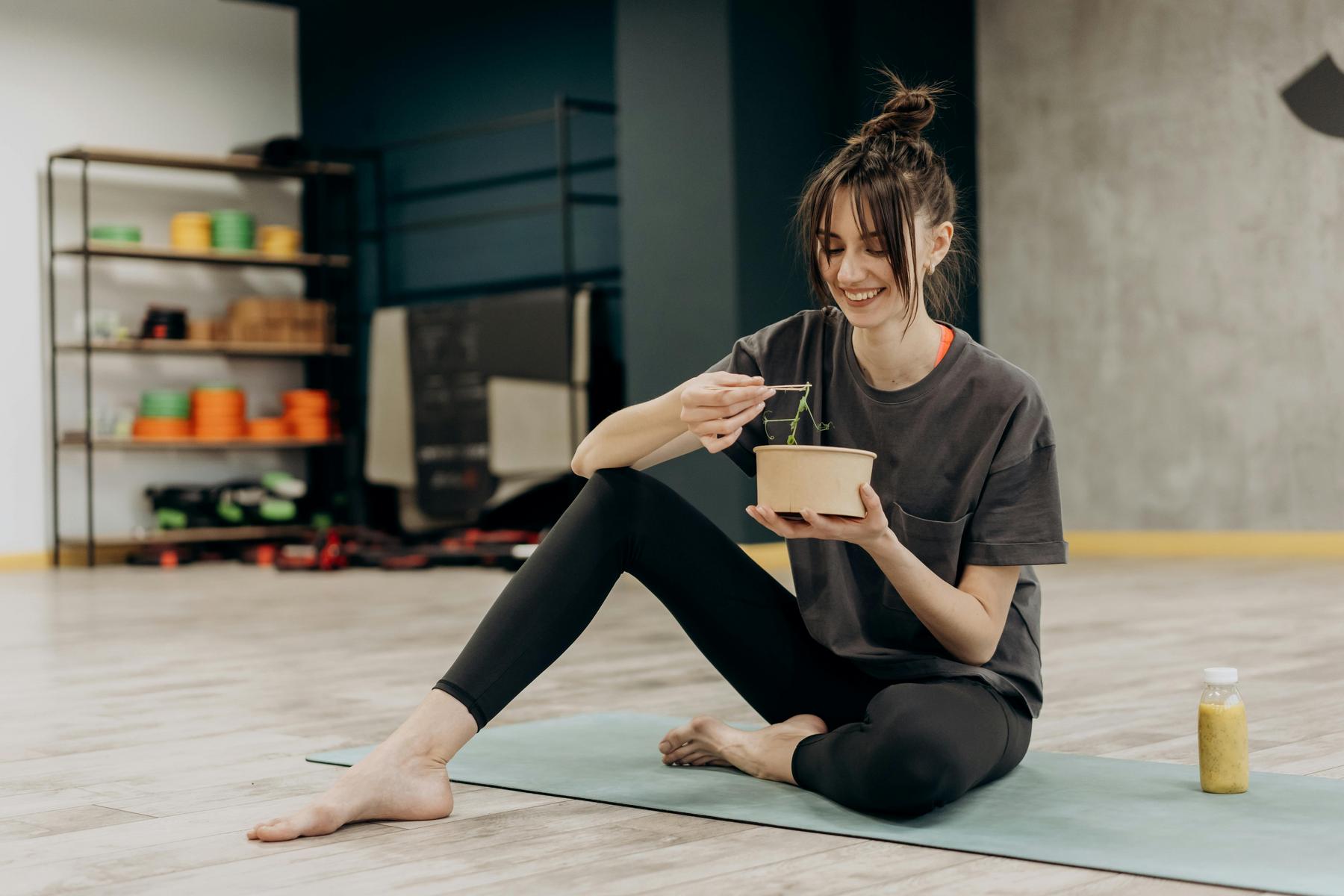 A woman sits cross-legged on a yoga mat indoors, eating from a bowl with a fork. A bottle of juice is placed beside her on the mat.