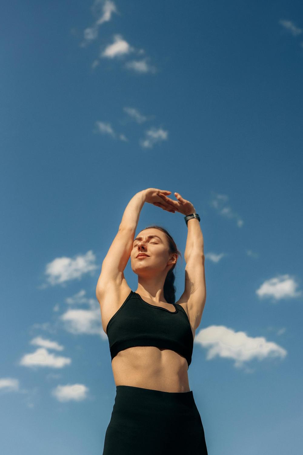 Woman in black sportswear stretching with arms raised overhead, standing outdoors under a blue sky with scattered clouds.