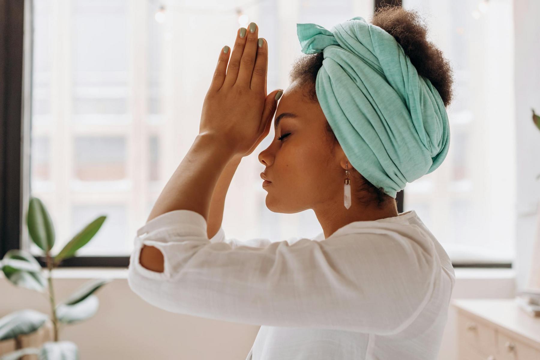 A person wearing a teal headscarf and white top holds their hands in prayer against their forehead, eyes closed, indoors near a window with plants.