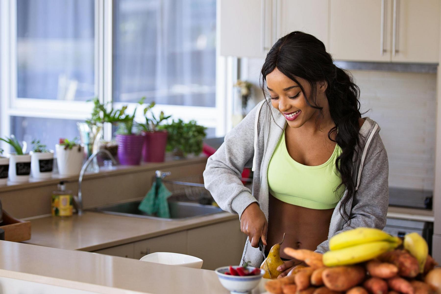 A woman in athletic wear smiles while slicing a pear in a kitchen with fruit and potted plants on the counter.