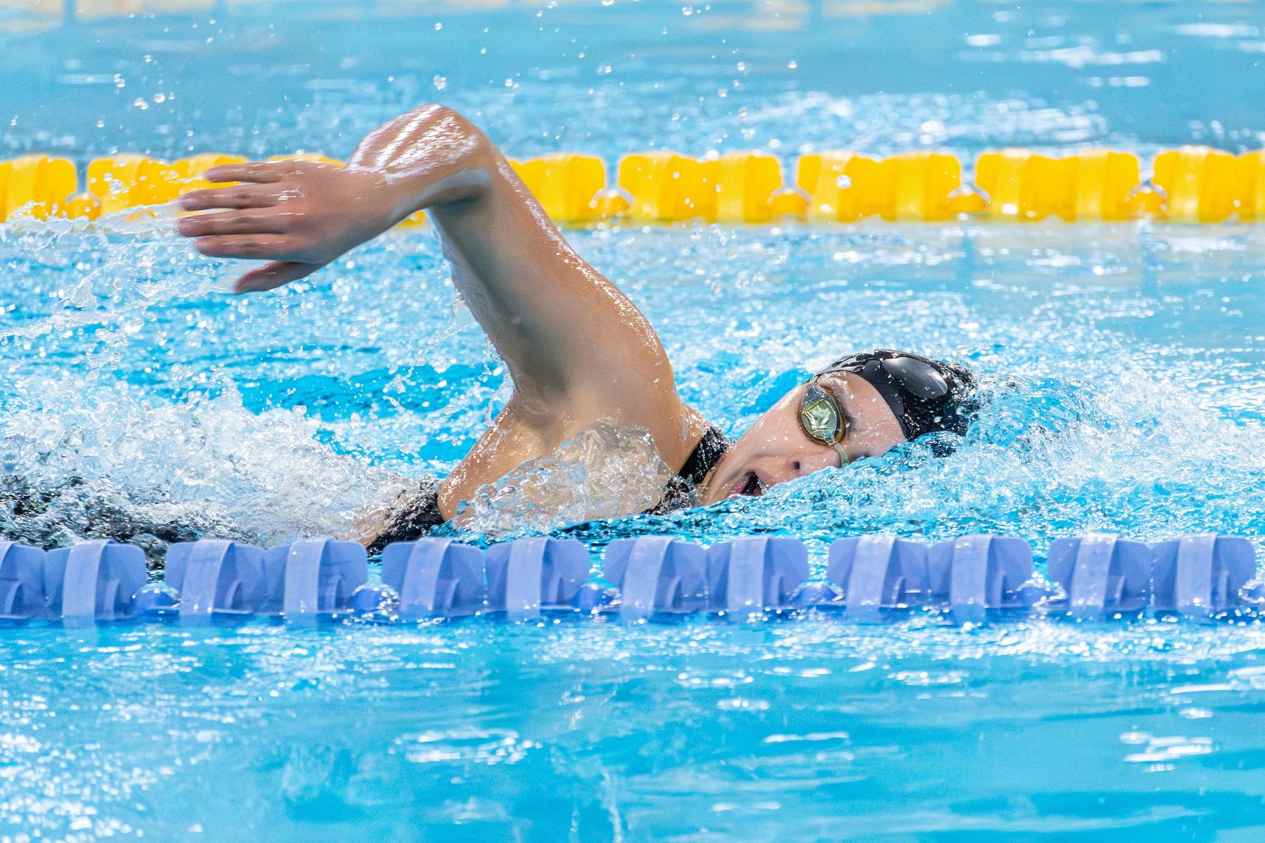 A swimmer wearing goggles and a swim cap performs freestyle stroke in a lane of an indoor swimming pool.