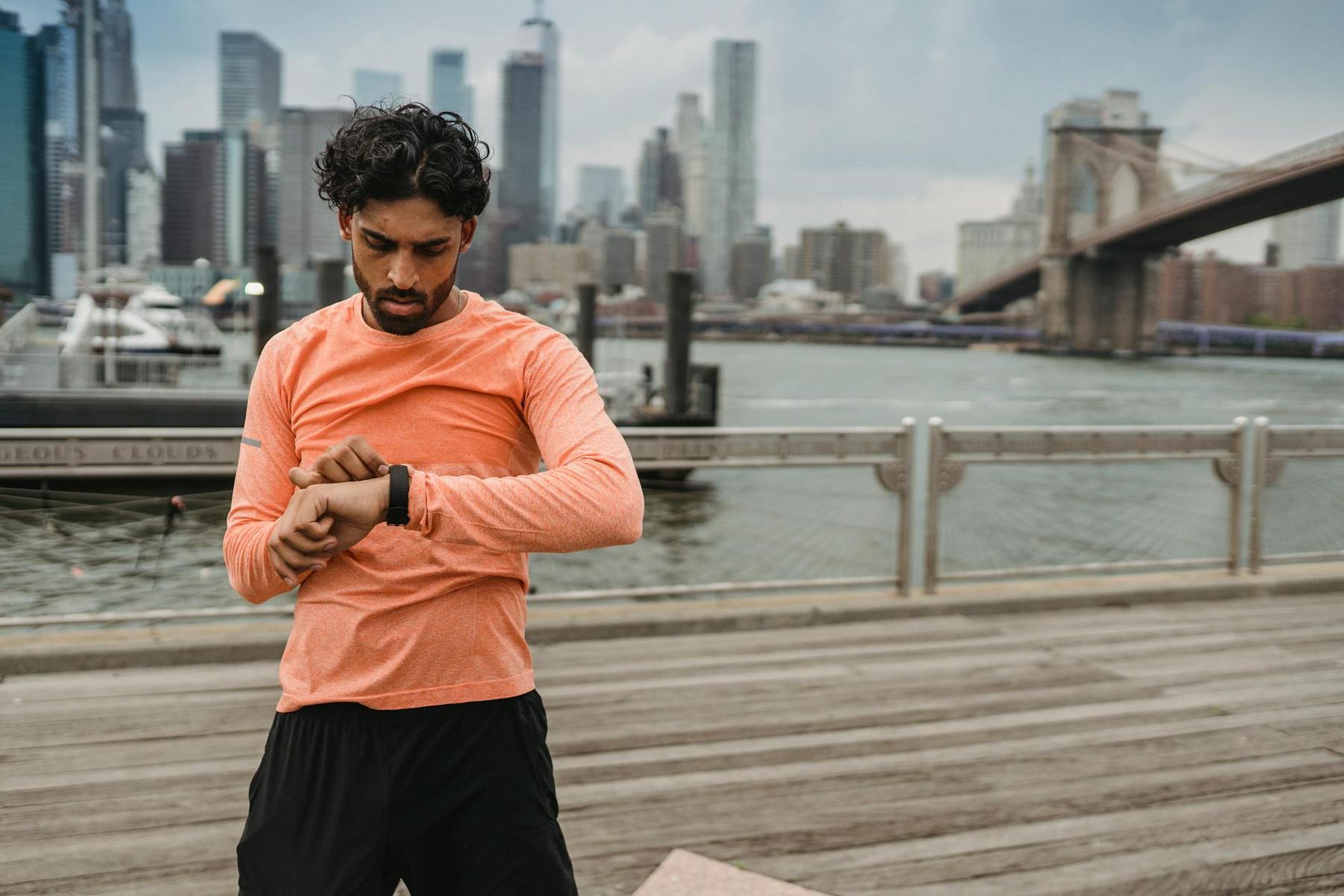 A man in athletic wear checks his smartwatch on a boardwalk by the river with a city skyline and bridge in the background.