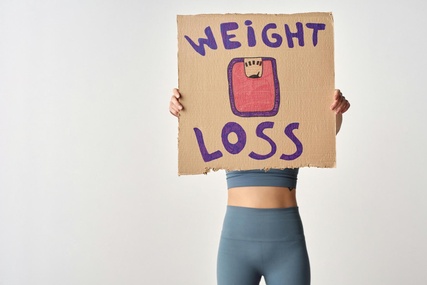 Person in athletic wear holds a cardboard sign reading "WEIGHT LOSS" with a drawing of a bathroom scale, obscuring their face.