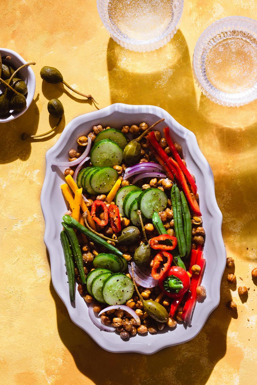 A white platter with sliced cucumbers, red and yellow bell peppers, green beans, capers, red onion, chickpeas, and jalapeños, set on a yellow surface with glass cups and a bowl of capers nearby.