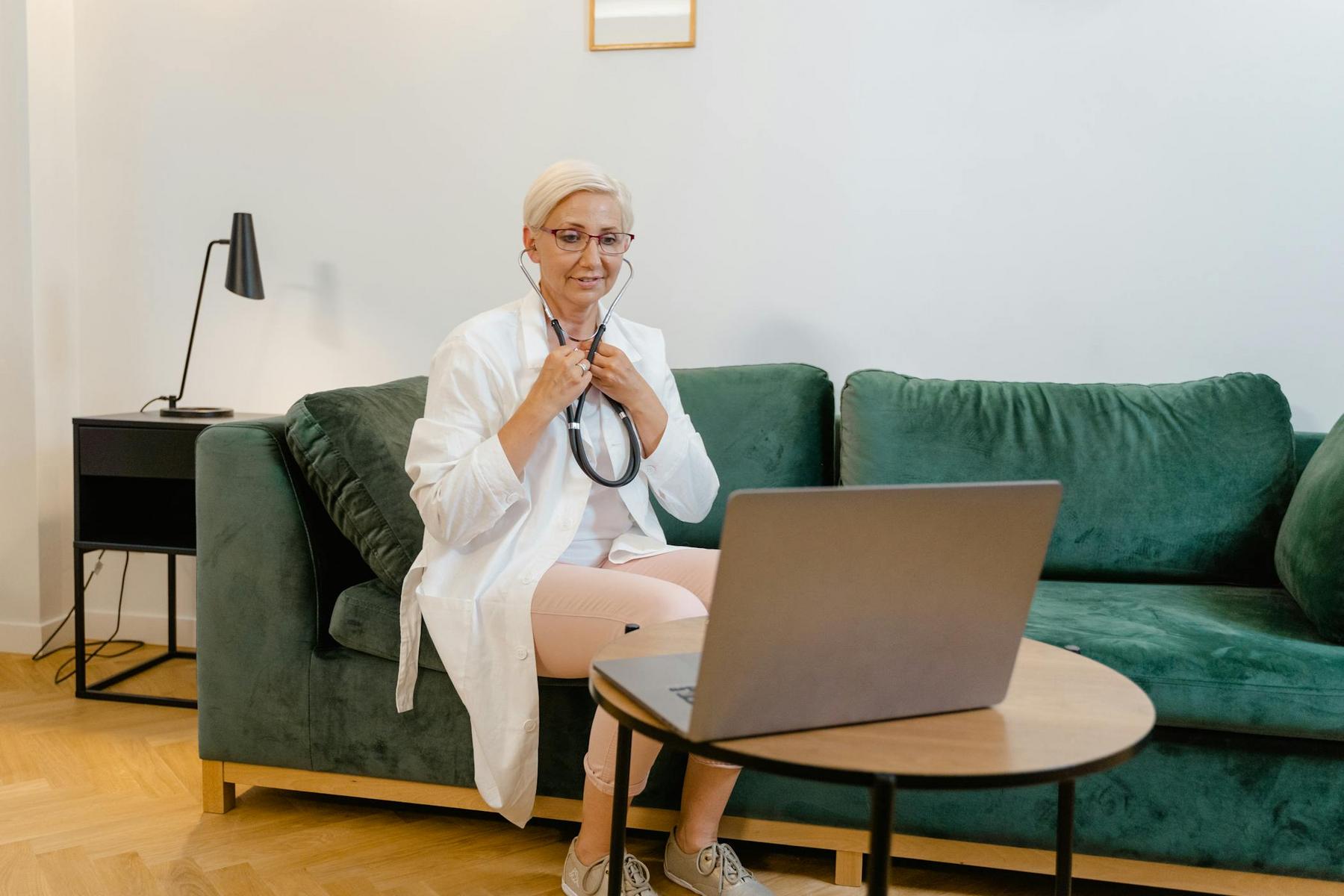 A doctor in a white coat sits on a green couch holding a stethoscope, looking at a laptop on a coffee table.