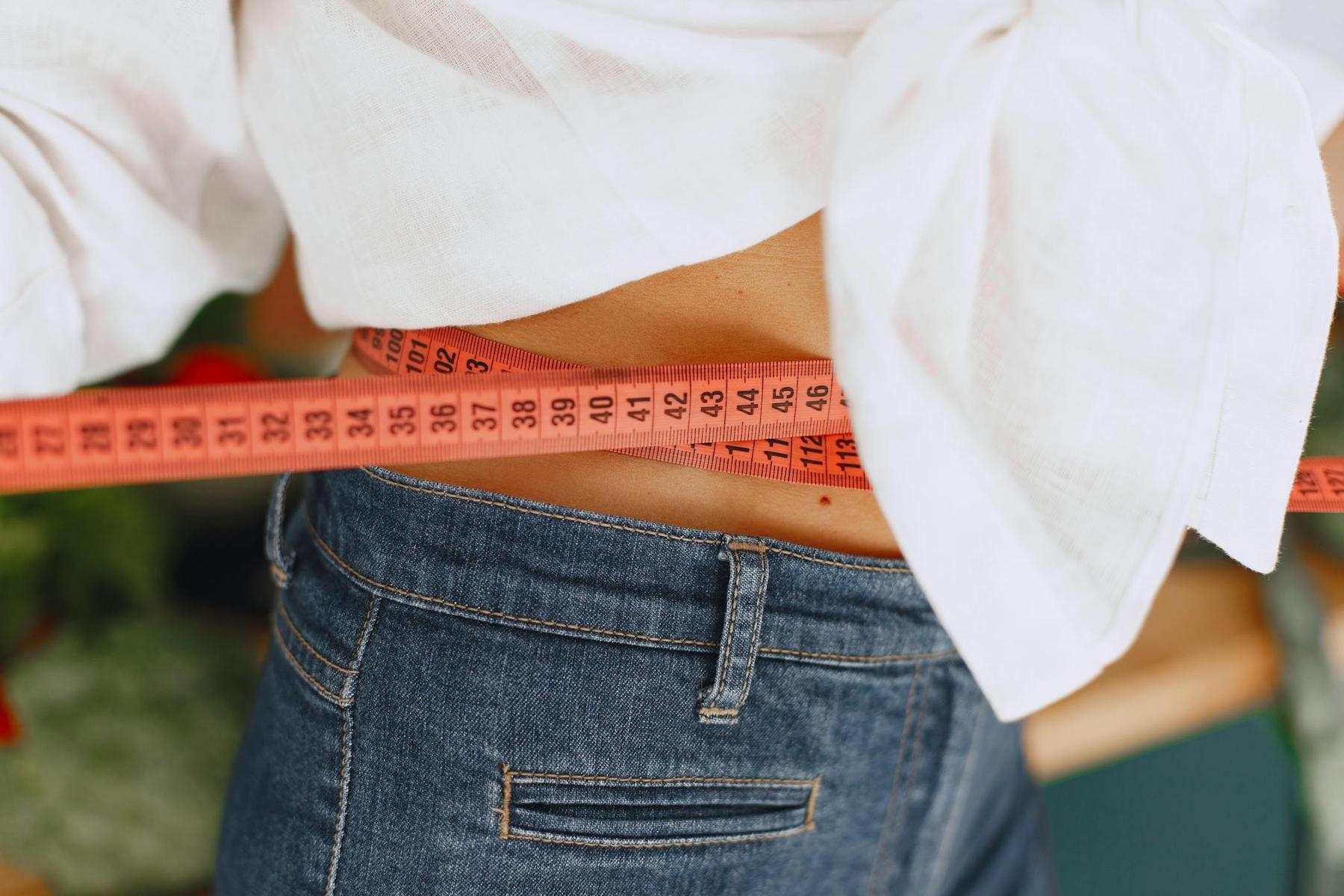 A person in jeans and a white shirt measures their waist with a red measuring tape.