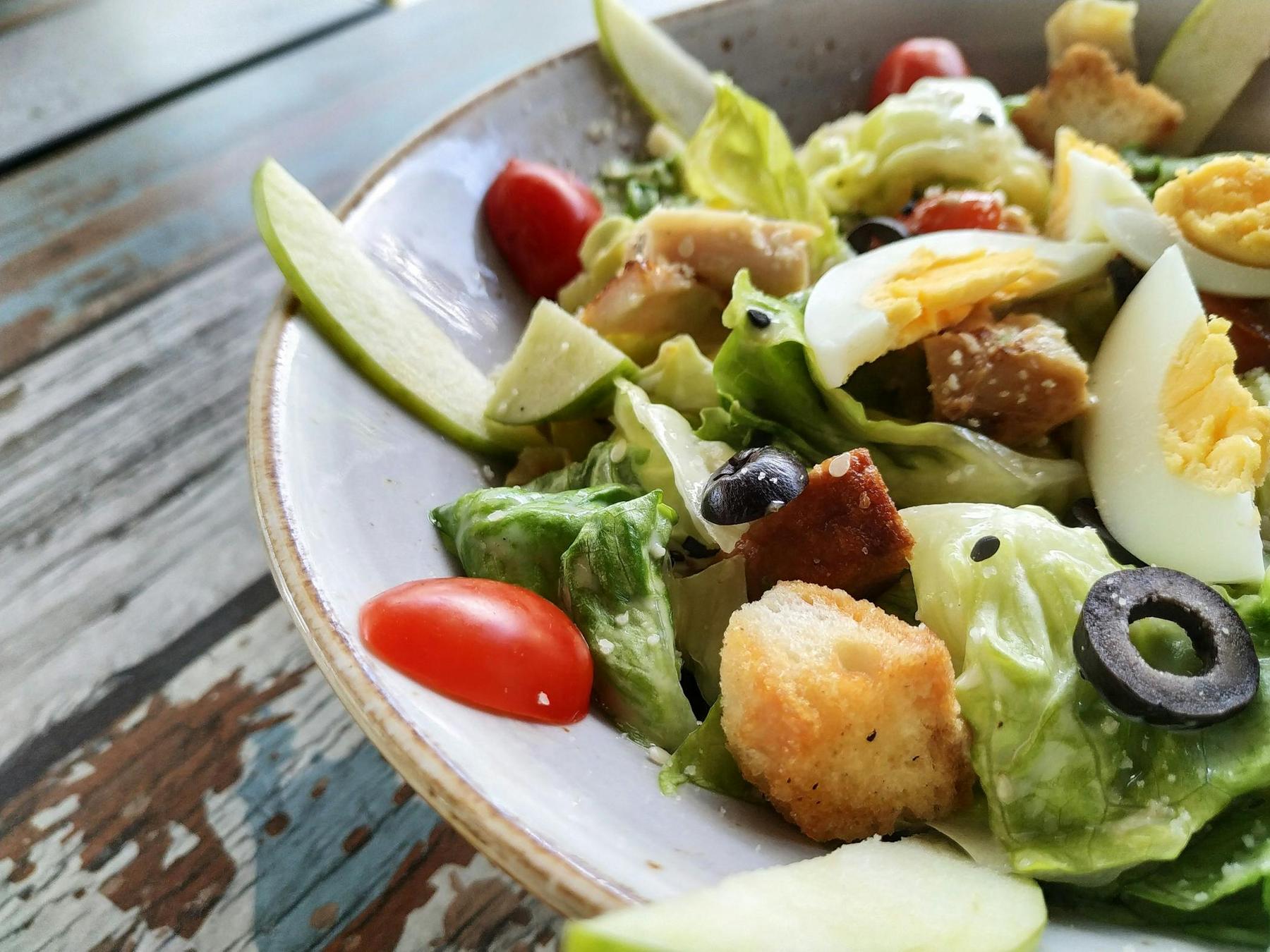 A close-up of a salad in a white bowl with lettuce, cherry tomatoes, croutons, black olives, hard-boiled egg slices, and green apple wedges on a rustic wooden table.