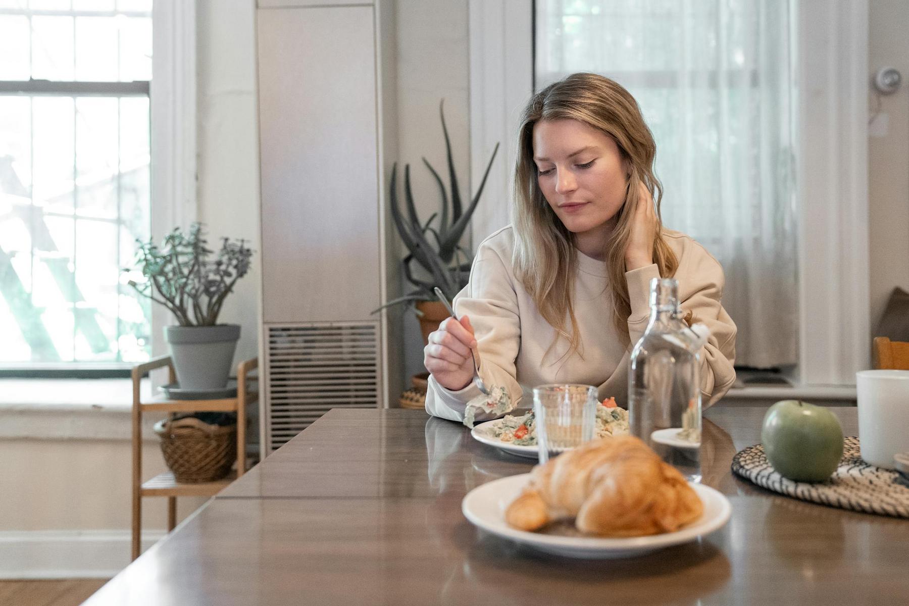 A woman sits at a dining table eating a meal, with a croissant, green apple, and a glass water bottle also on the table. Houseplants and large windows are in the background.