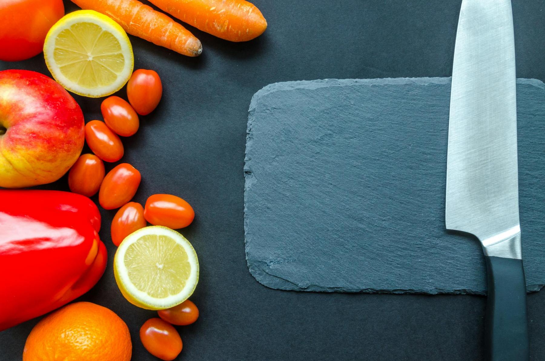 Vegetables, fruits, a lemon, and a knife are arranged on a dark surface with a slate cutting board in the center.