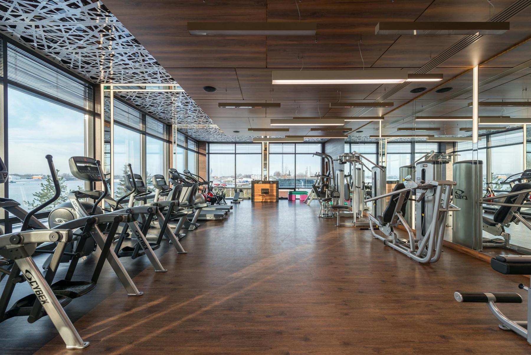 Modern gym with cardio and strength equipment, large windows, wooden floors, mirrored walls, and a patterned ceiling.