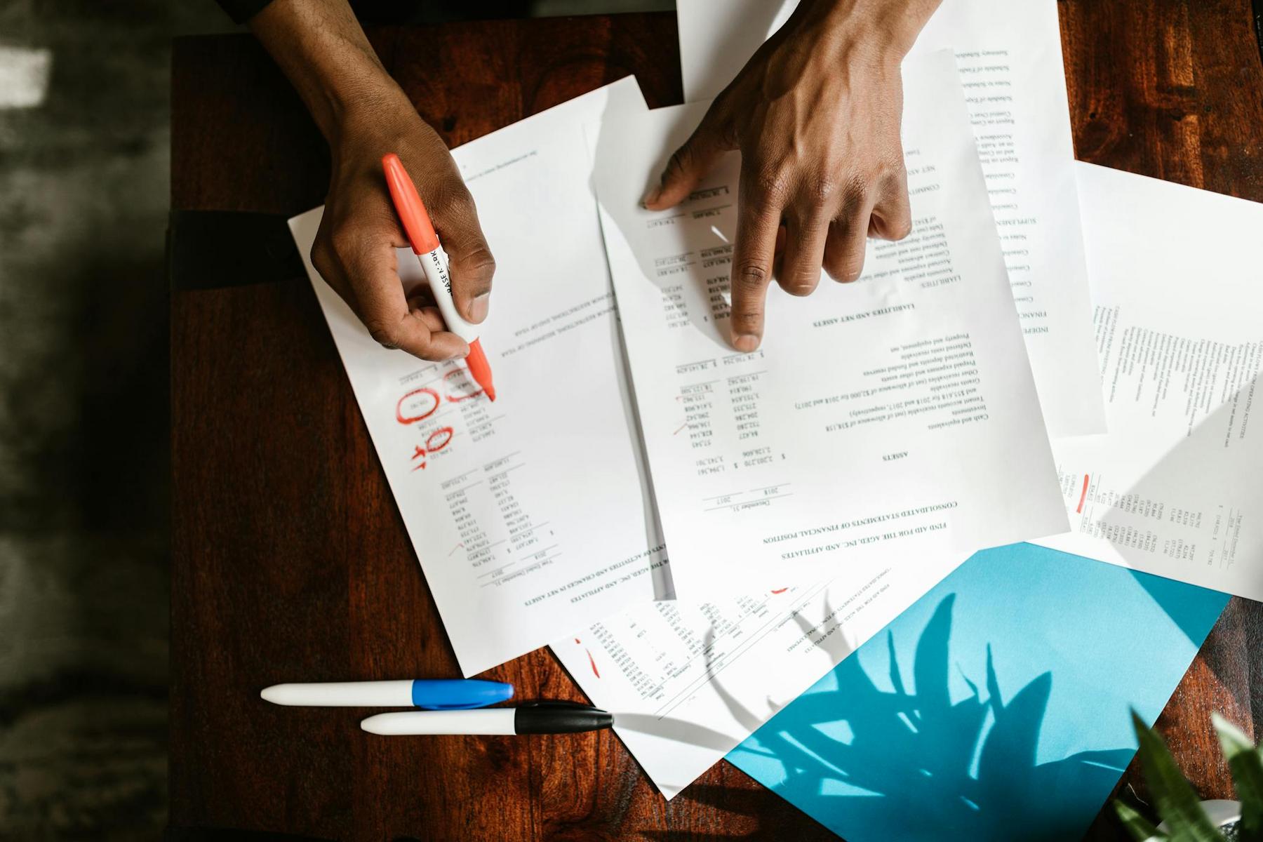 Hands reviewing and marking documents with a red pen on a wooden desk, alongside blue and black pens and a blue folder.