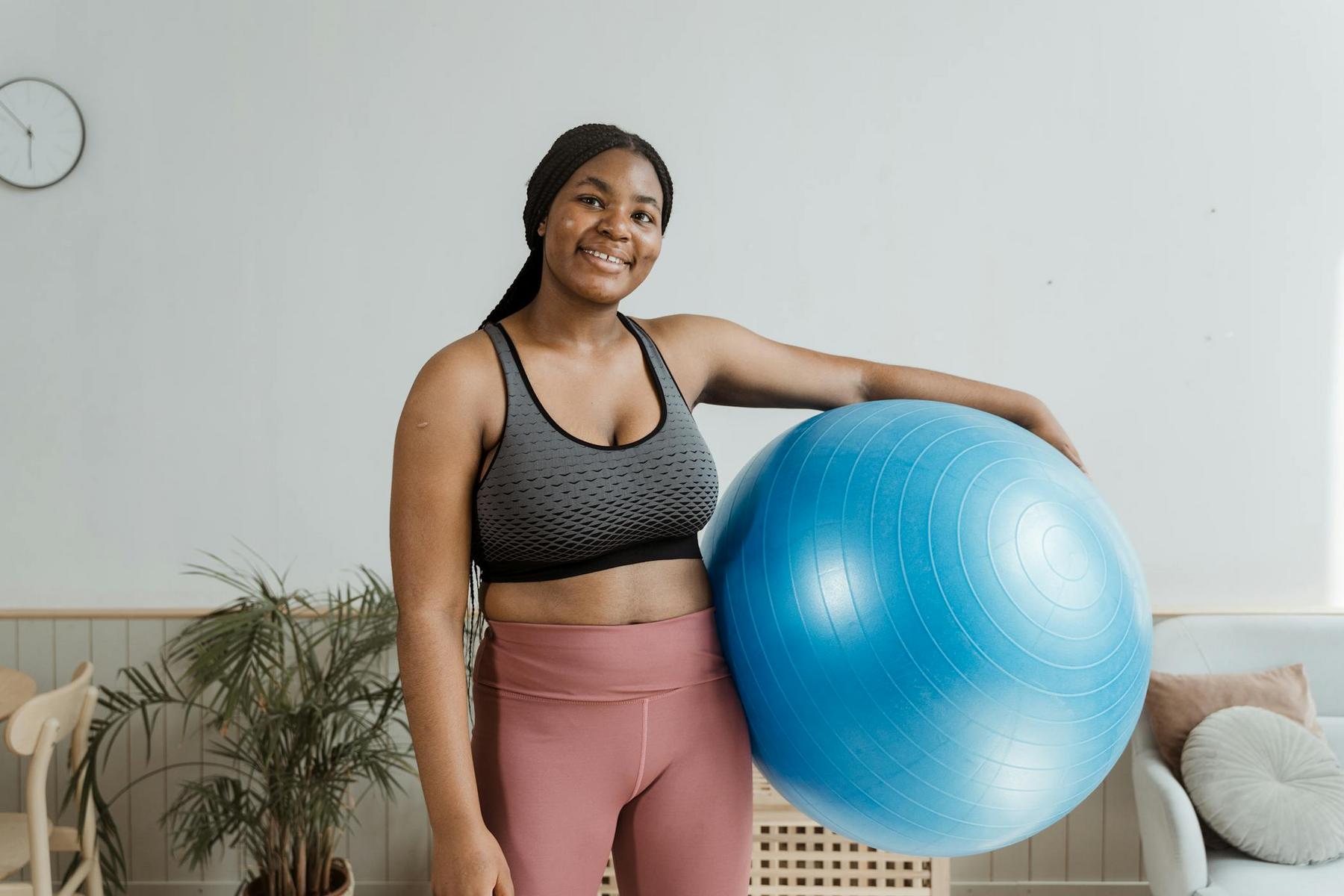 A woman in athletic wear stands indoors, smiling and holding a large blue exercise ball under her arm.