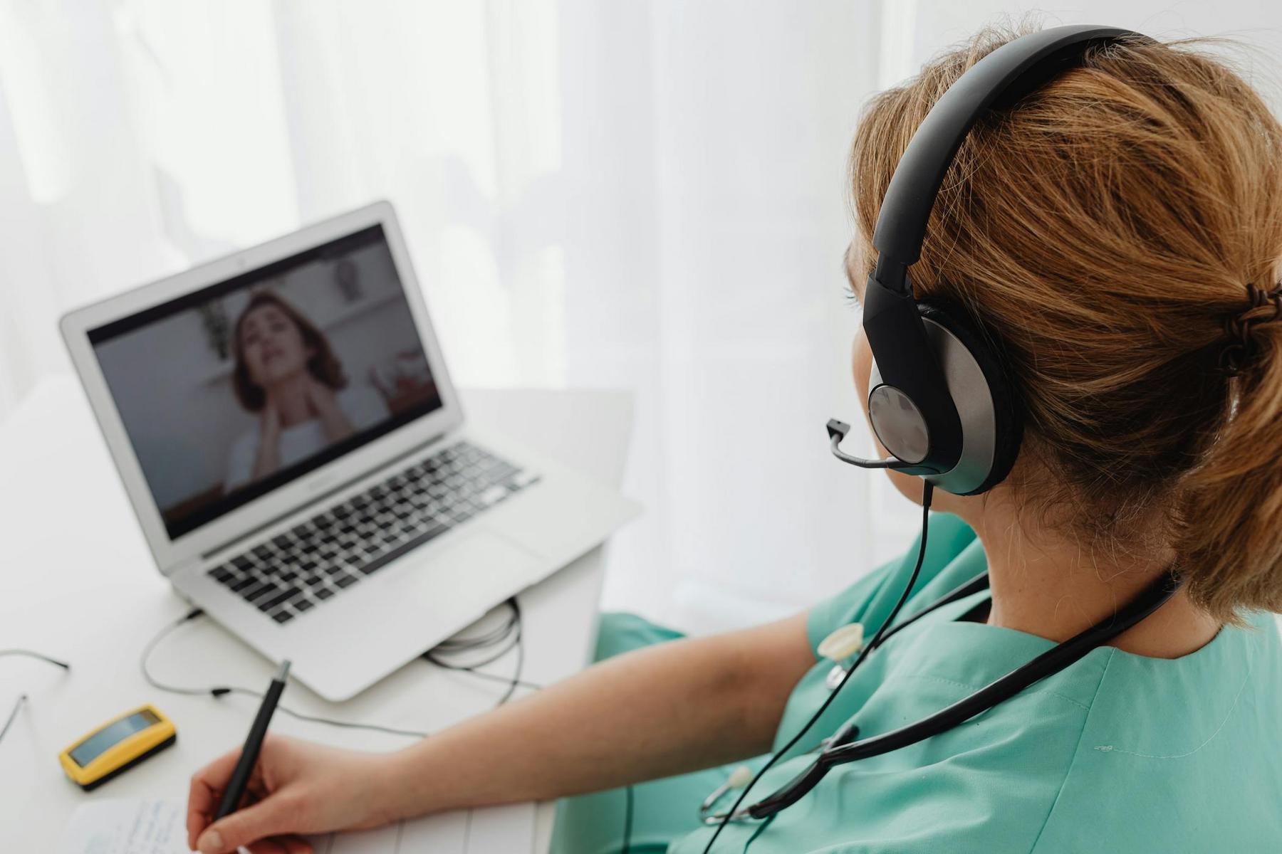 A healthcare professional wearing a headset consults with a patient on a video call, taking notes at a desk with medical equipment nearby.