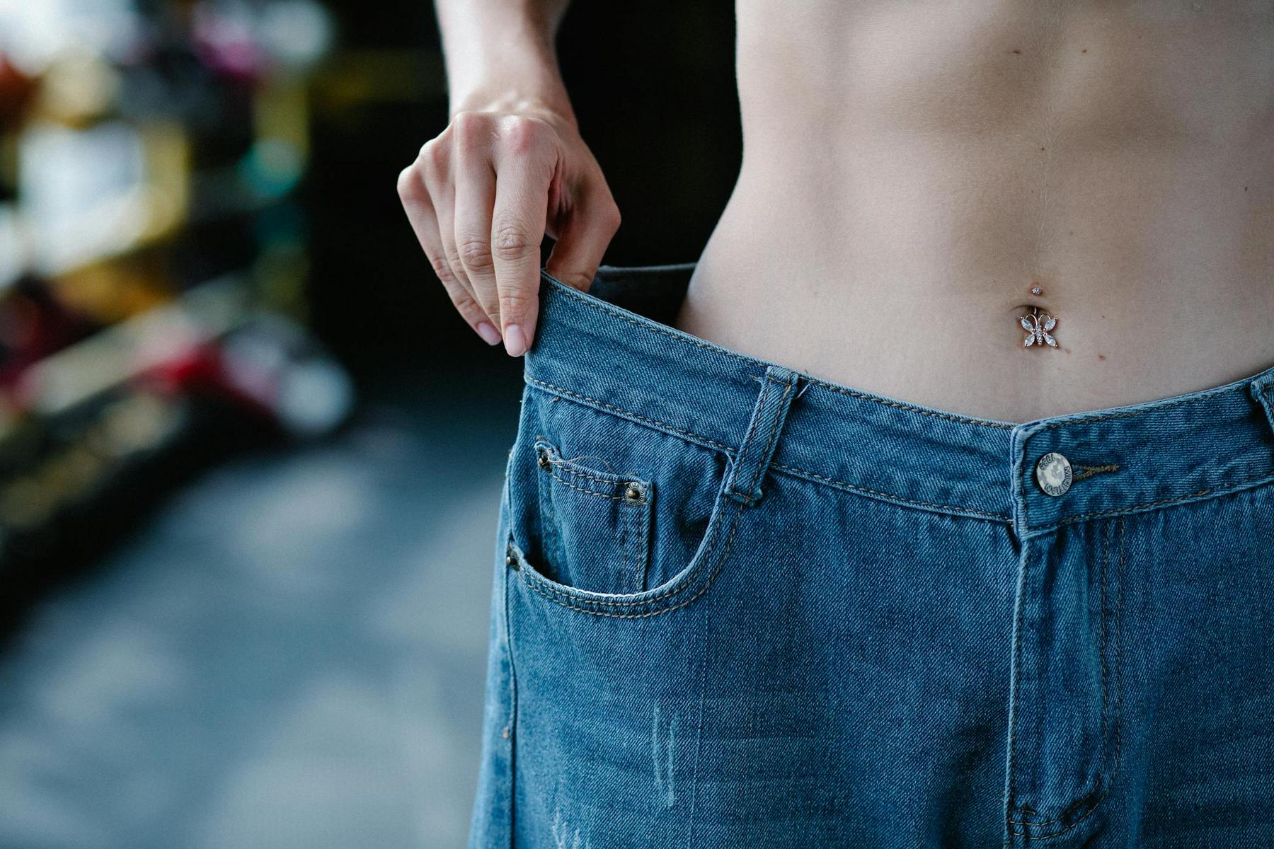 Person showing weight loss by wearing loose jeans, pulling the waistband away from their stomach in a dimly lit setting.