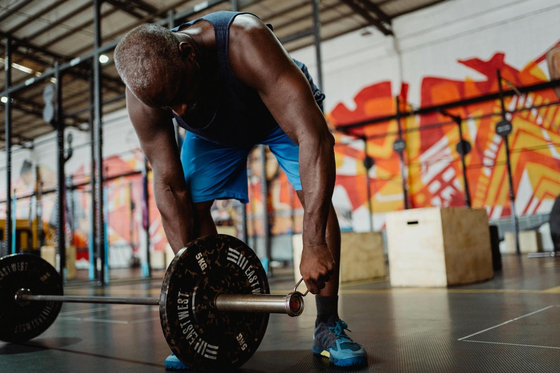 A man in athletic wear bends down to grip a barbell in a gym with colorful murals and wooden boxes in the background.