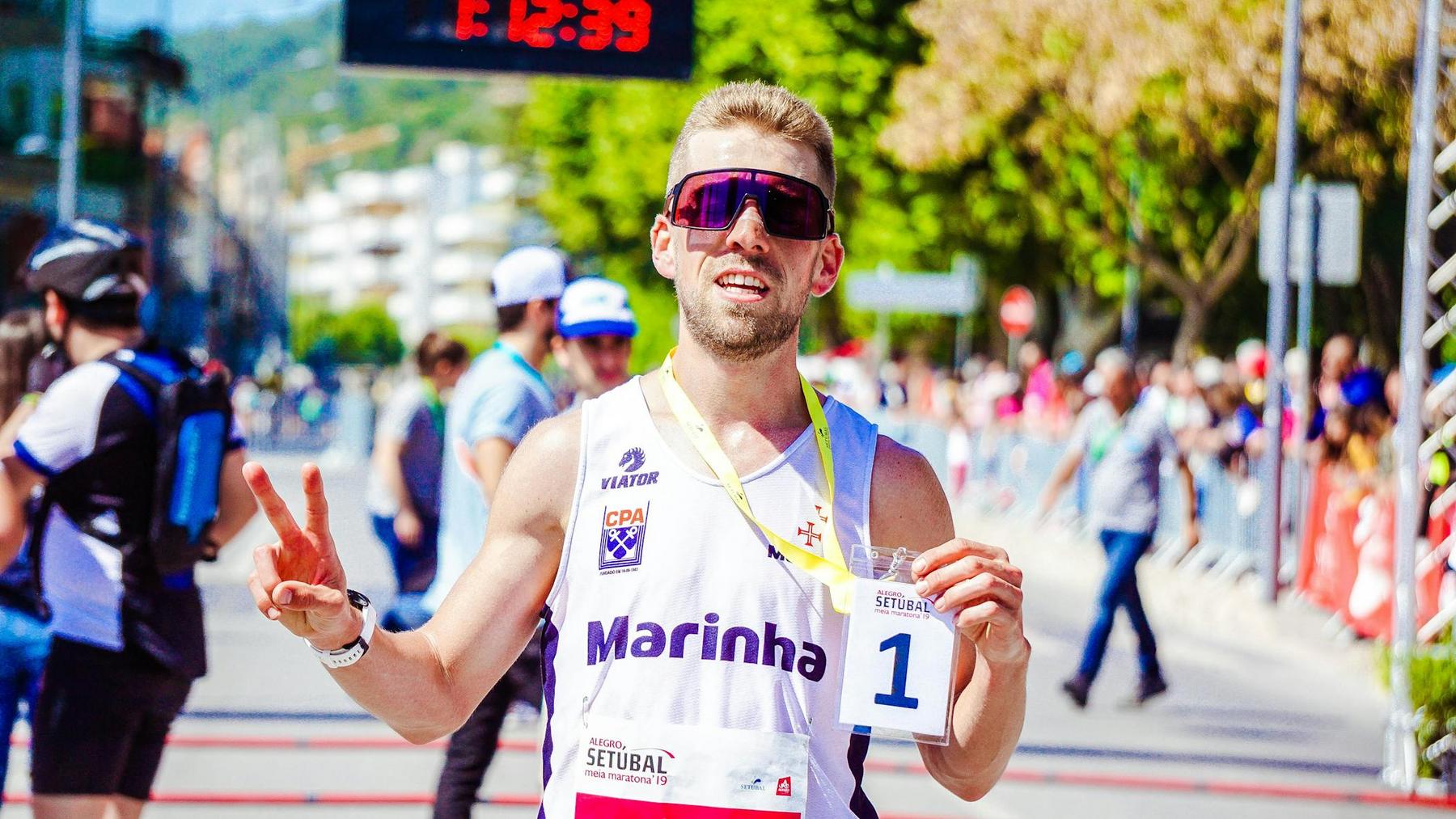 A male runner wearing sunglasses and holding a medal and a race bib numbered 1 poses with a peace sign at a finish line during a daytime race event.