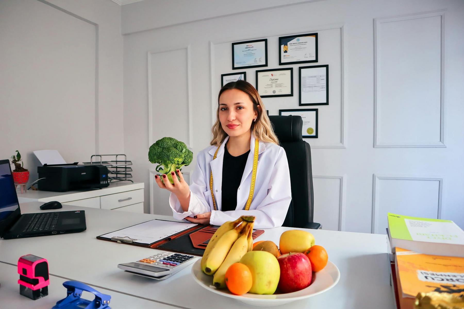 A person in a white coat sits at a desk, holding a broccoli. The desk has a laptop, calculator, and a bowl of fruit. Certificates hang on the wall in the background.