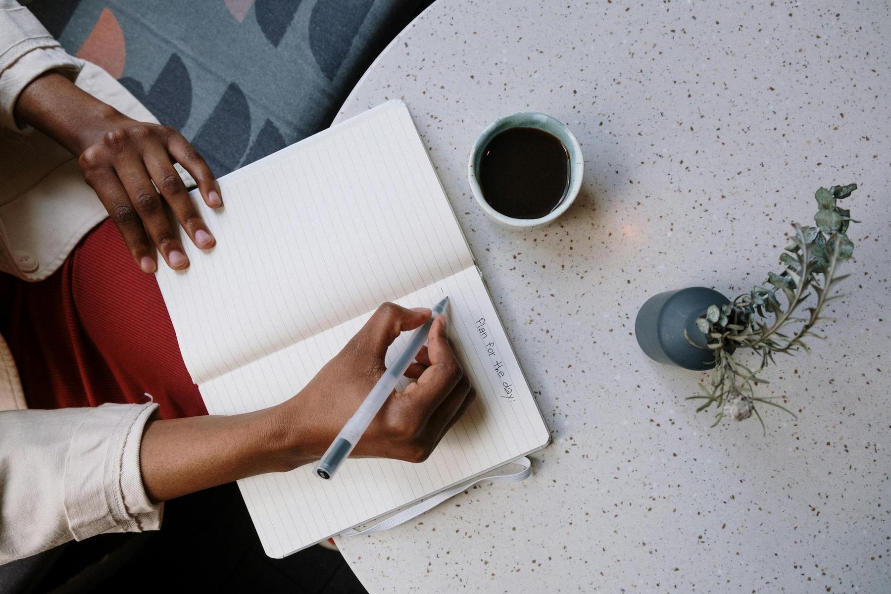 A person writes "Time for the day" in a notebook at a round table with a cup of coffee and a small potted plant.