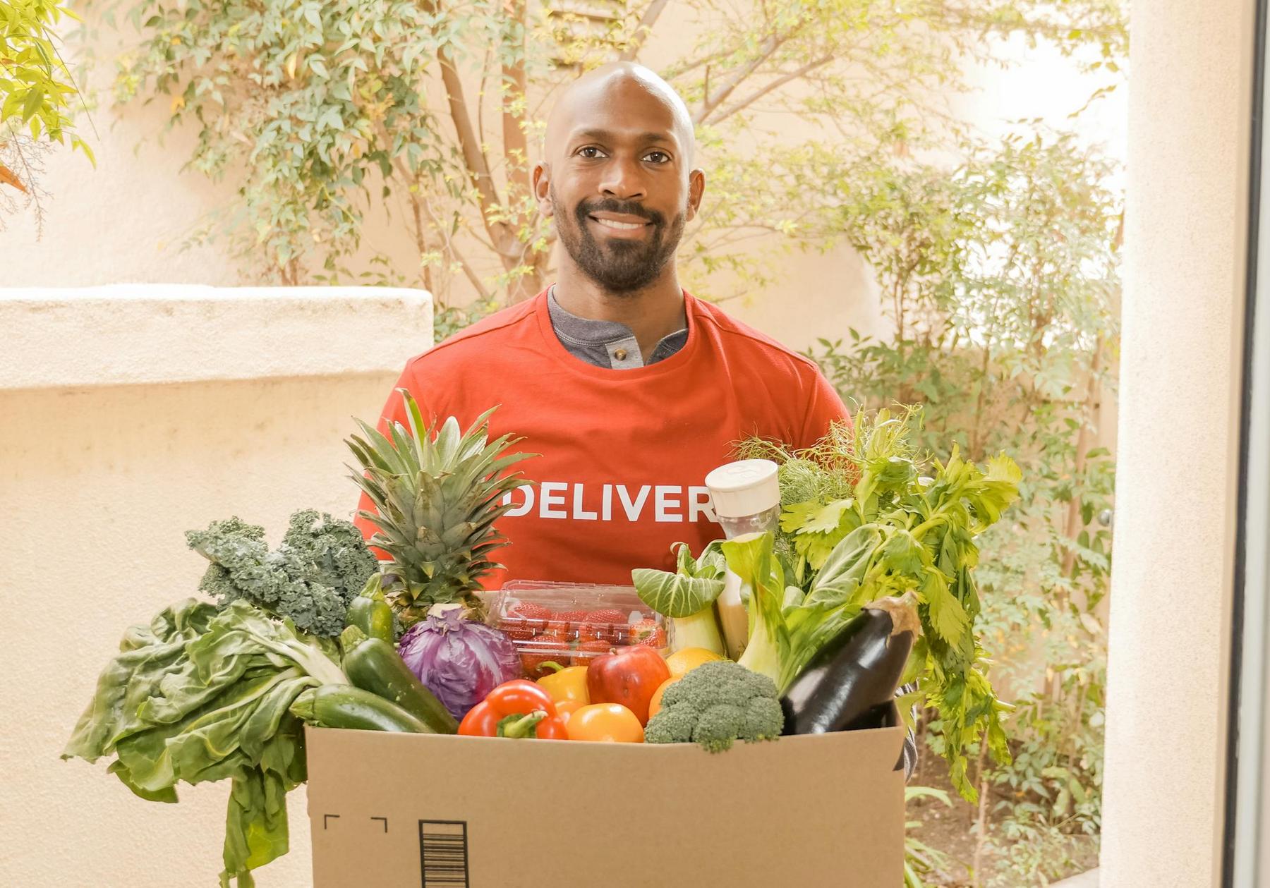 A man wearing a red delivery shirt stands outside holding a box filled with assorted fresh vegetables and fruits.