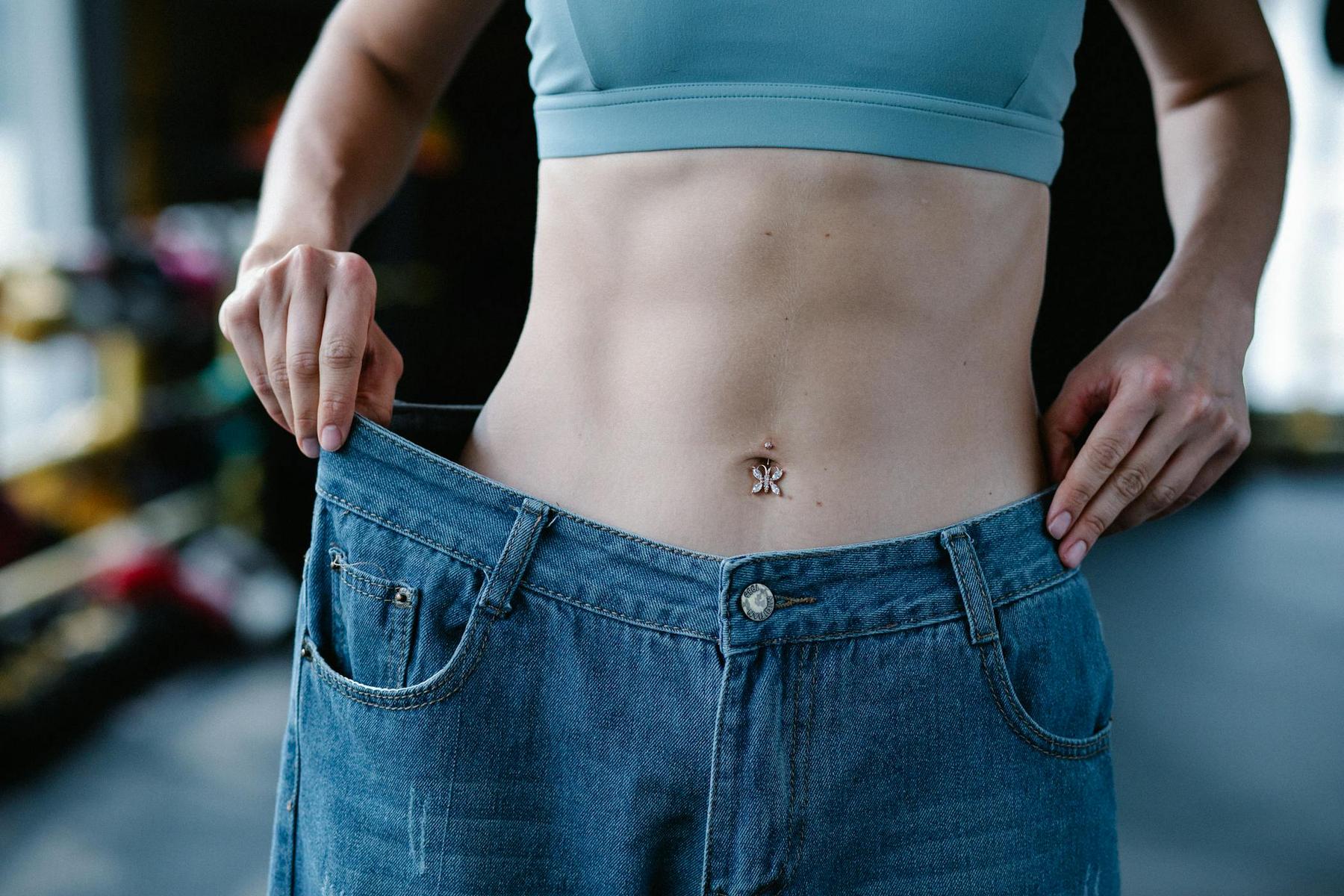 Person in a sports bra holds out oversized jeans, highlighting significant weight loss.