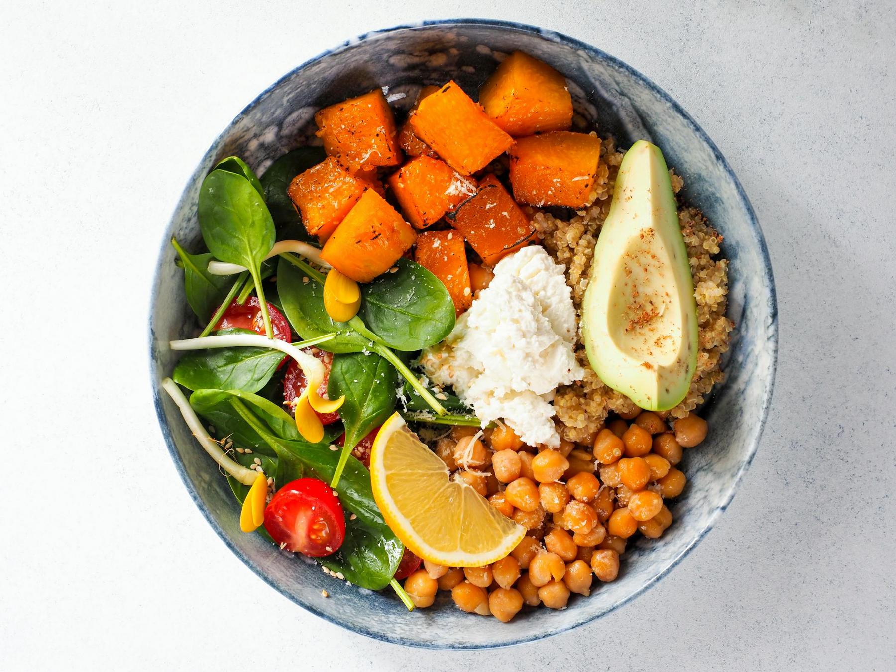 A bowl with roasted squash, avocado, quinoa, chickpeas, ricotta cheese, spinach, cherry tomatoes, sunflower sprouts, and a lemon wedge on a light surface.