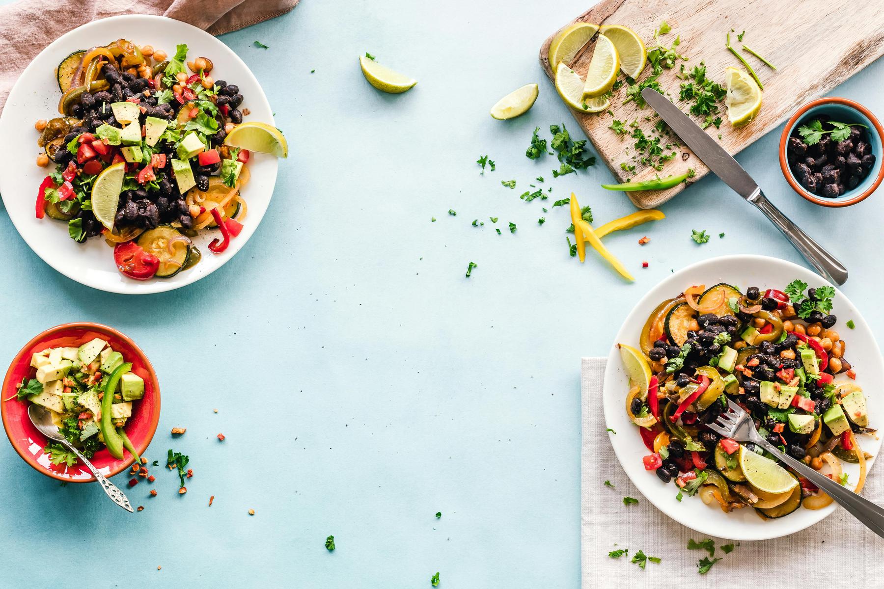 Two plates of salad with black beans, avocado, and vegetables, surrounded by lime wedges, herbs, and a bowl of salad on a blue surface. A cutting board with a knife and fork is visible.