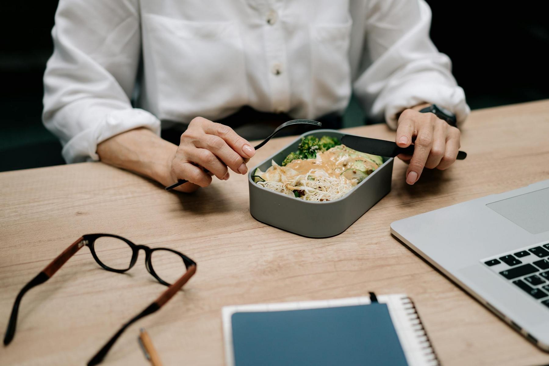 Person in a white shirt eating noodles and vegetables from a lunch container at a desk with a laptop, glasses, and notebook.