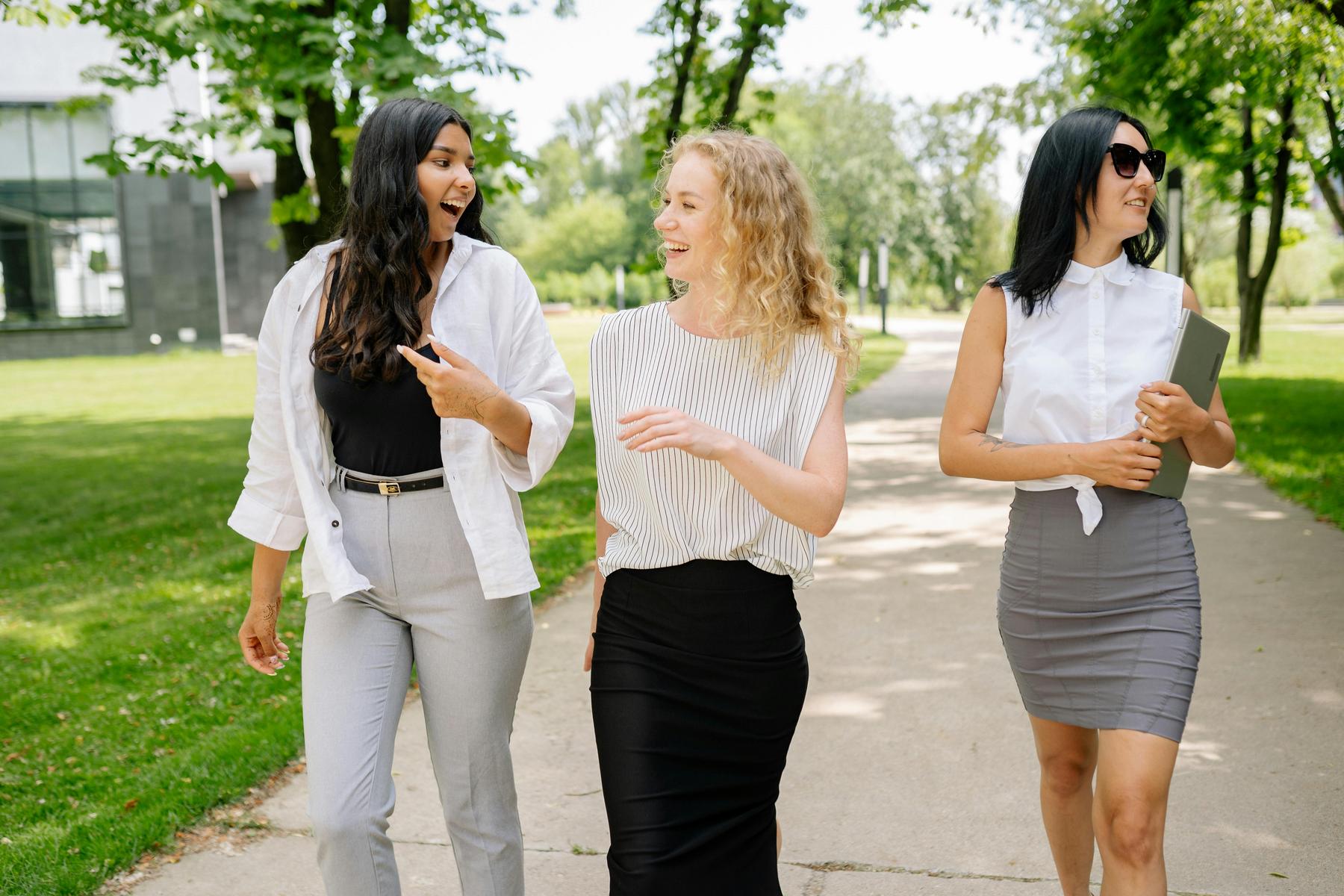 Three women in business attire walk and talk together on a tree-lined outdoor path, with one holding a tablet and the others engaged in conversation.