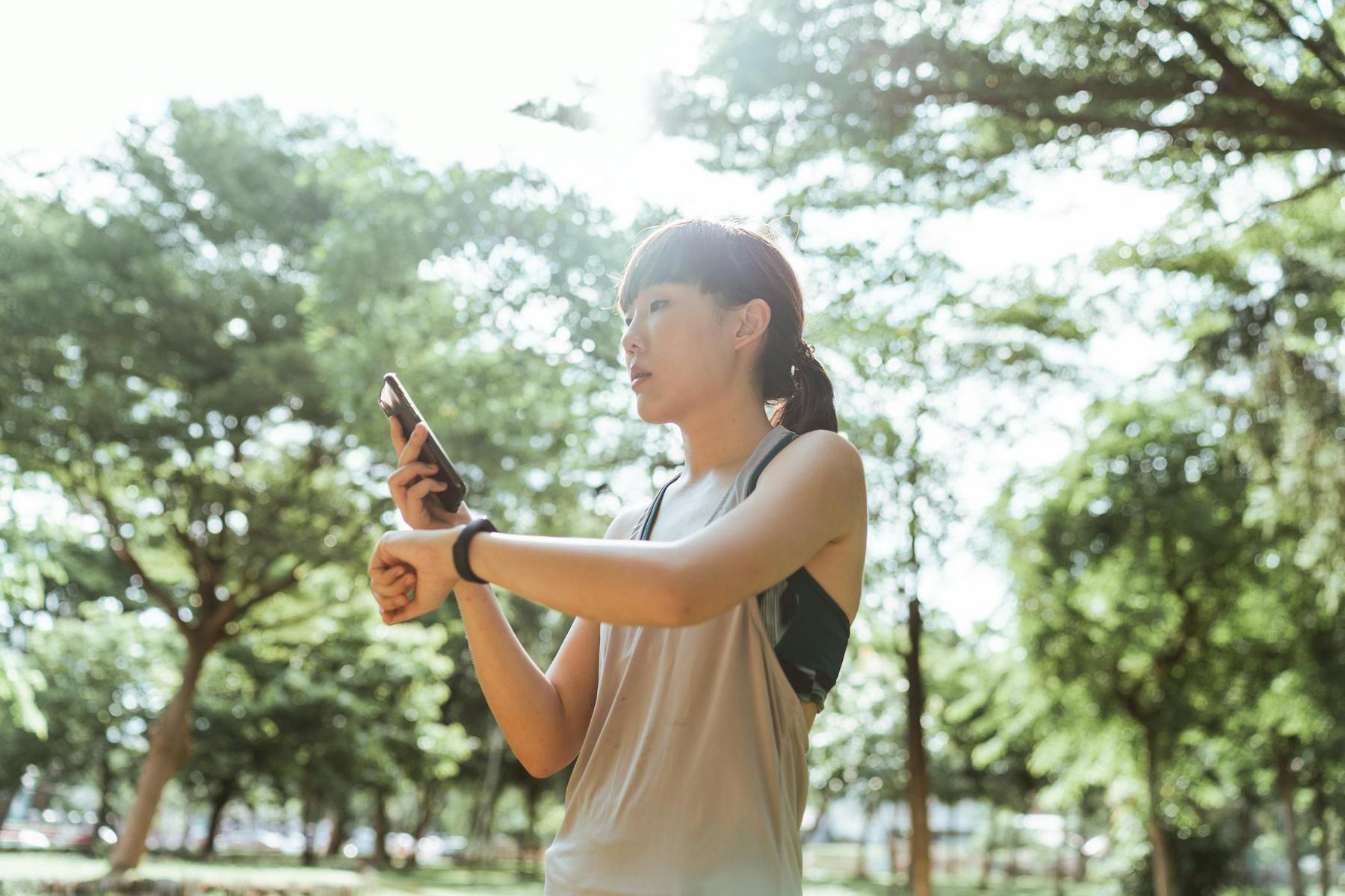 A person in athletic wear stands outdoors in a park, looking at a smartwatch on their wrist and holding a smartphone in their other hand.