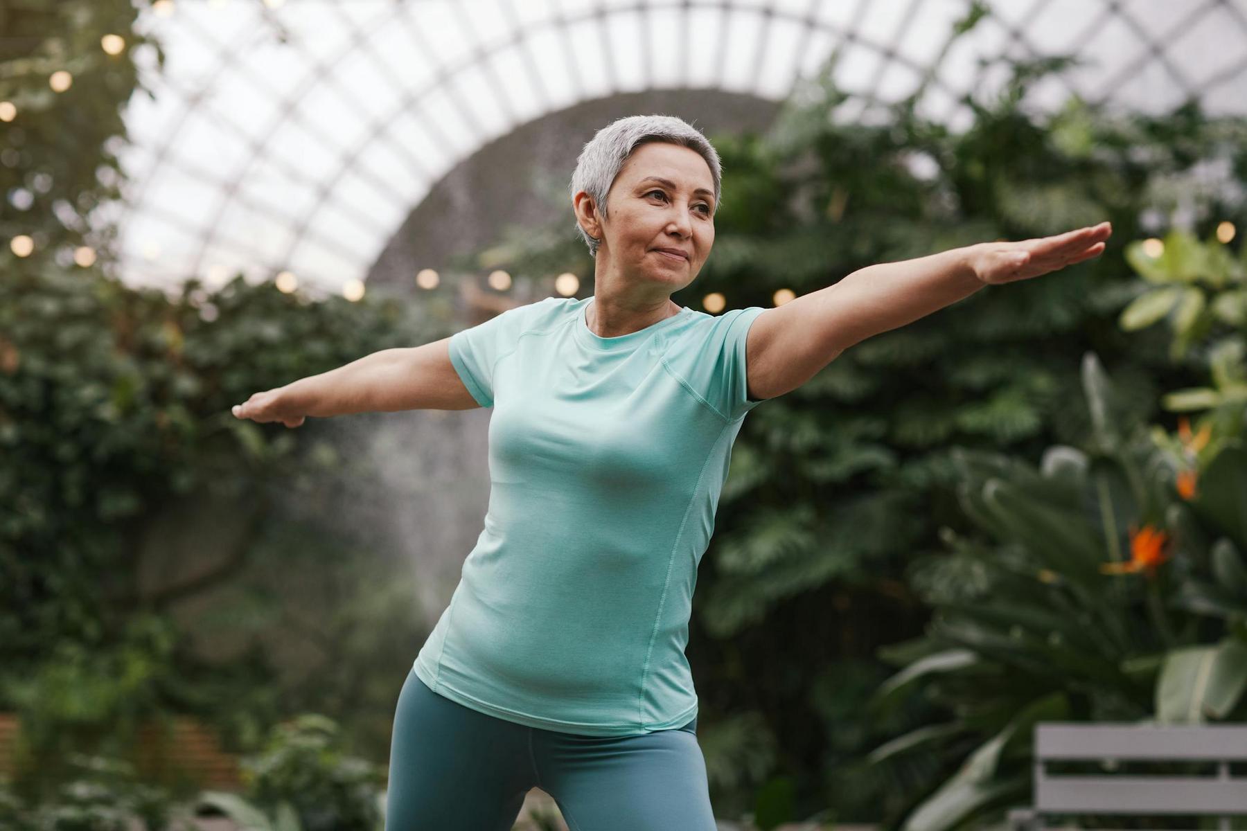 A woman with short gray hair practices a yoga pose in a garden setting, wearing a teal shirt and leggings.