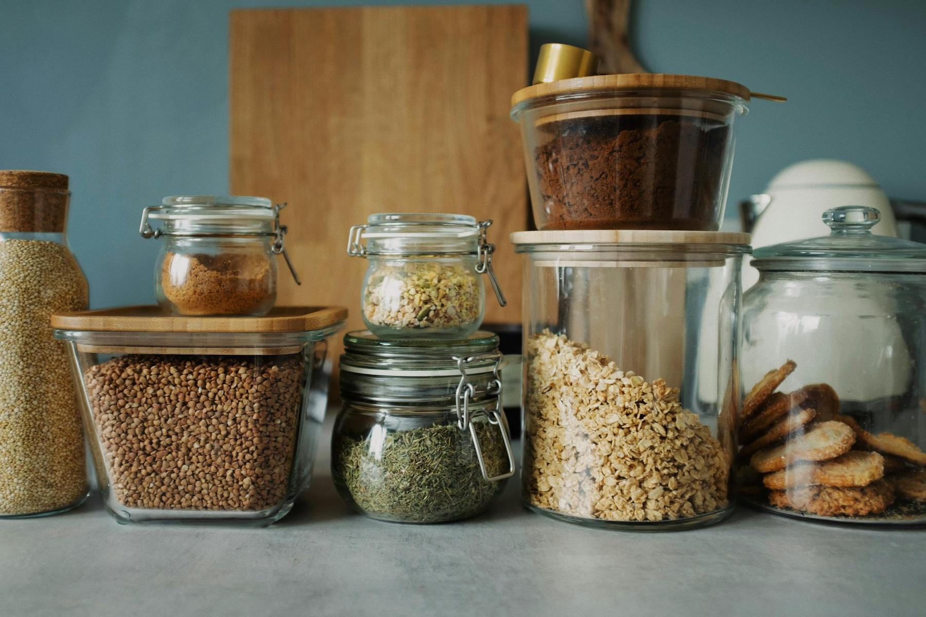 Assorted glass jars with bamboo lids contain various dried foods like grains, herbs, oats, and cookies, neatly arranged on a kitchen counter. A wooden cutting board is in the background.