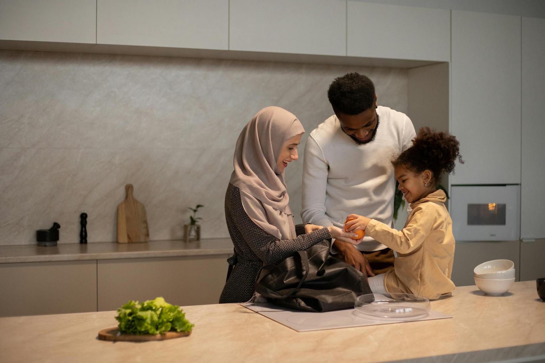 A family of three stands in a modern kitchen. A woman in a hijab and a man assist a young girl who is unpeeling an orange. A cutting board with lettuce is nearby.