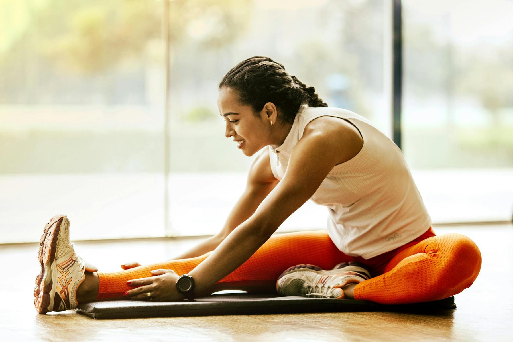 A woman wearing a white sleeveless top and orange leggings sits on a yoga mat indoors, stretching forward to touch her foot.