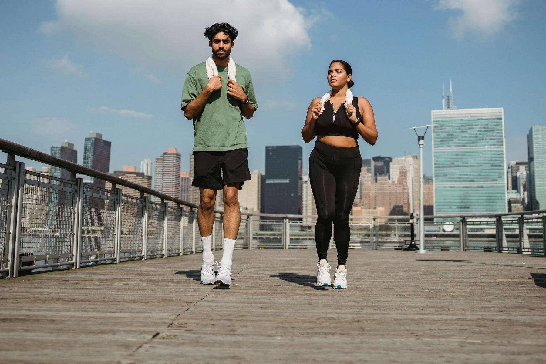 Two people walk on a wooden boardwalk with towels around their necks, wearing athletic clothing, with a city skyline in the background.