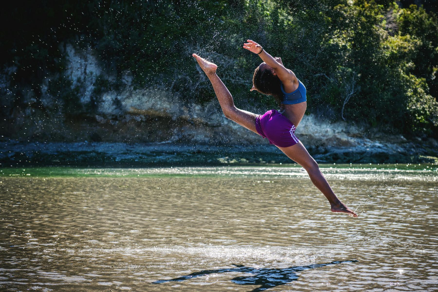 Person in athletic clothing performing a high jump with an arched back and extended arms and legs over shallow water outdoors.